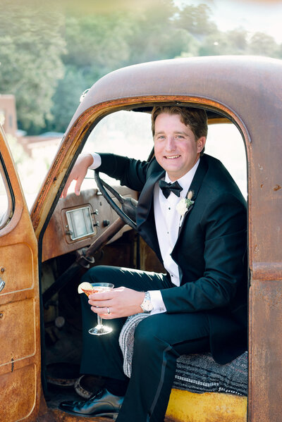 Groom in tuxedo sits in vintage pick up truck while drinking cocktail