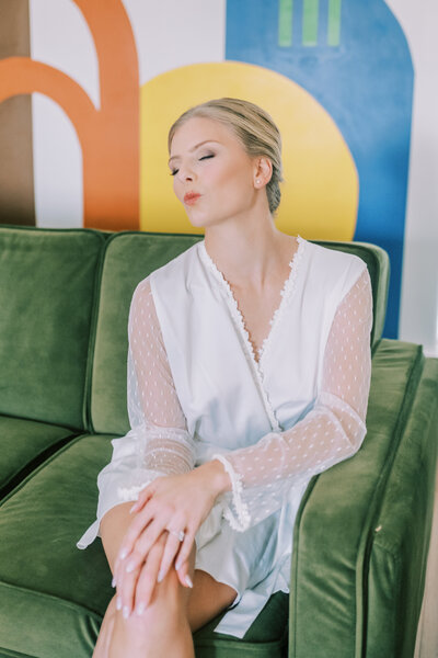 Bride in a white robe posing on a green couch while getting ready for her wedding