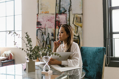 The image captures a contemplative moment of a woman sitting at a glass table, seemingly deep in thought or reading. She has her hand to her chin and is holding what appears to be a magazine or catalog. The woman is wearing a casual white sweater, and her hair is styled down. Behind her is a colorful abstract painting, adding an artistic touch to the room. Natural light filters through the window to her left, and there are various plants around, which contributes to a tranquil and creative atmosphere. A teal blue velvet chair sits next to her, indicating a stylish interior design.