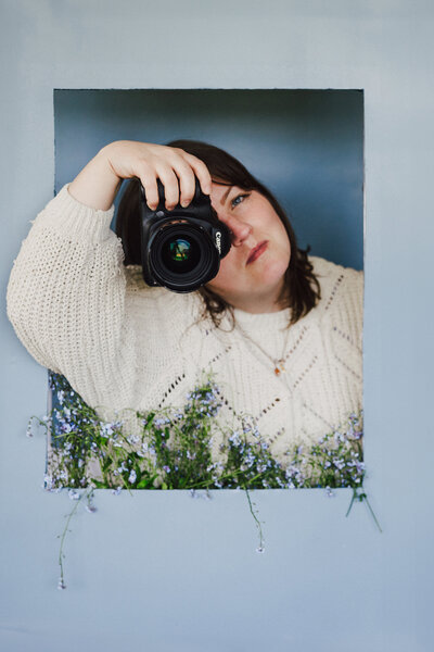 Artistic self portrait of Dani Keith. She is in a blue frame with blue wildflowers. A camera is covering half of her face.