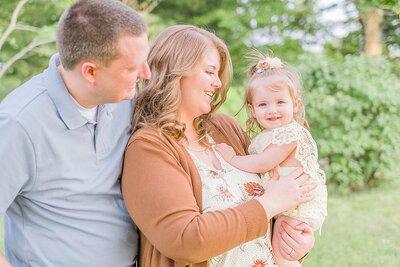 A mom and dad looking at their little girl and smiling. Girl is smiling right at camera