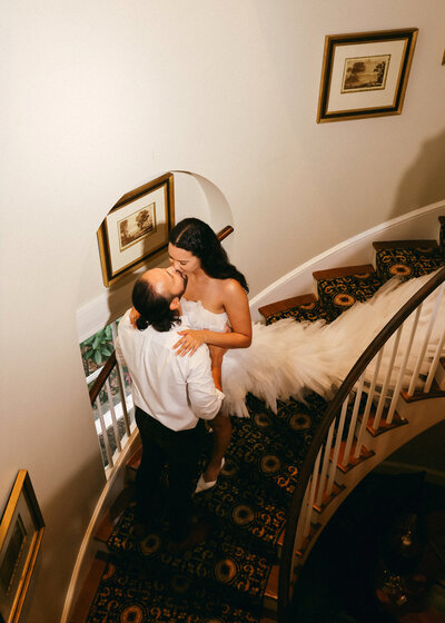 Kaitland and Kramer walk hand-in-hand down the aisle inside Boxwood Estate. The couple is smiling, with Kaitland wearing a flowing white gown and Kramer in a sharp suit. The venue features beautiful chandeliers and soft lighting, adding a romantic ambiance to the setting. Rows of white chairs are neatly arranged on either side of the aisle, and floral arrangements decorate the space, creating an intimate and sophisticated atmosphere.