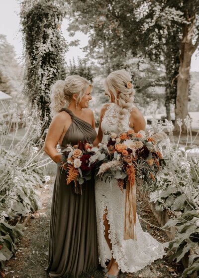 A bride and bridesmaid with boho hair and floral bouquets smiling at each other.