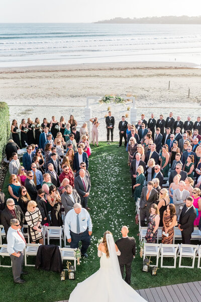 A stunning aerial view of a beachside ceremony at Newport Beach House in Middletown, RI. The bride walks down the aisle with waves rolling in the background and guests seated along a manicured lawn path.