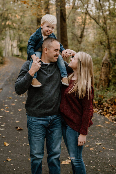 Mom wearing red shirt and jeans looking at her husband and smiling who is wearing a gray sweater and jeans  with young son on his shoulders sticking his tongue out at Patapsco State Park in Maryland