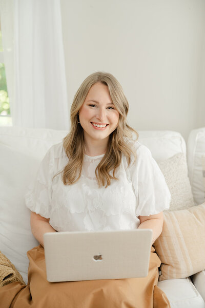 Woman in a white top and burnt orange skirt seated at a white couch holding her Macbook Pro and smiling