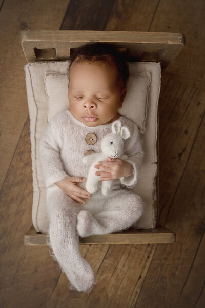 newborn boy laying in tiny wooden bed, holding stuffed bunny during hamilton, ON newborn photoshoot
