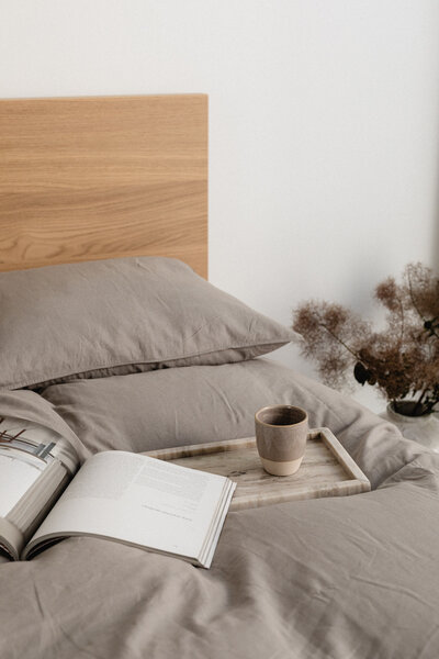 A beautifully styled bed featuring soft grey linen bedding, a wooden headboard, and a tray holding an open book and a warm mug. A vase with dried flowers sits beside the bed, adding a rustic yet refined touch. Created for Luxe Linens’ social media, this image evokes comfort, relaxation, and modern luxury.