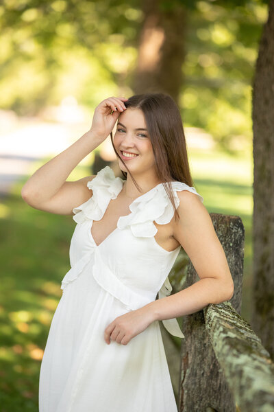 senior touching her hair as she stand in golden hour light in a white dress celebrating with her senior portraits