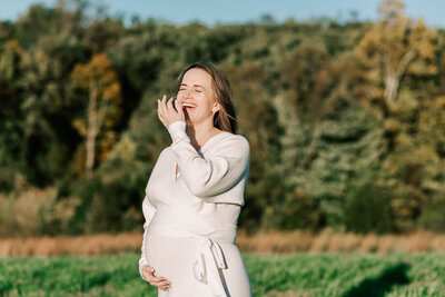 A pregnant mom in a white sweater dress holding her belly, standing in the fields of 48 Fields