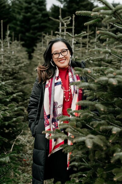 Woman standing among evergreen trees, smiling warmly in a red turtleneck sweater and colorful patterned scarf on a cool winter day.