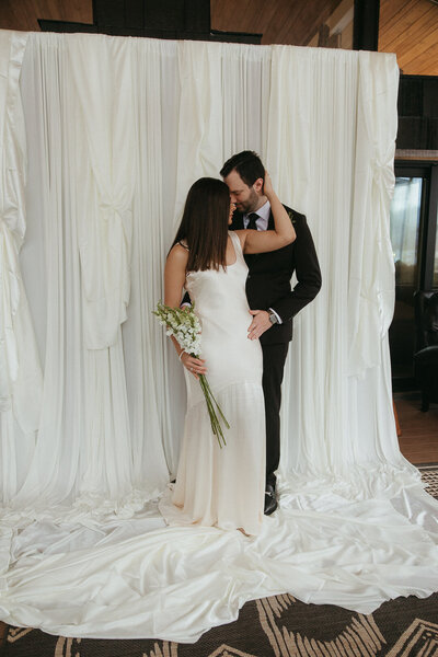 A finger lakes bride and groom cuddling in close in front of a trendy, ivory satin backdrop. Photo by Dash and Boheme Photography. 