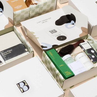 A collection of Bespoke Lane & Co. custom gift boxes, elegantly designed with minimalist patterns and branded logos. The boxes contain curated items such as skincare products and grooming accessories, reflecting a luxurious and personalized unboxing experience. The packaging features soft green checkered designs and sleek typography, emphasizing the brand's attention to detail and premium service.