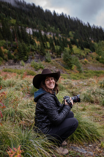 Smiling adventure photographer in a wide-brim hat kneels in an alpine meadow with a camera in hand.