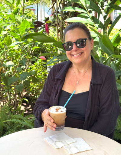 Woman smiling and sitting outside at table, holding iced coffee, and with tropical greenery behind her