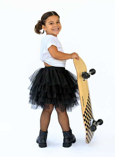 Little girl standing with a skateboard wearing a black tutu and fishnets. 