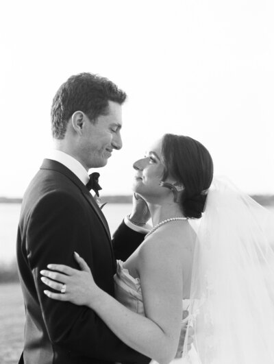 Black and white close-up photo of a bride and groom smiling at each other with water in the background.