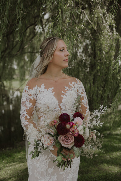 A portrait of a bride under a willow tree holding a colorful summer bouquet, from her backyard wedding in western new york. Photo by Dash and Boheme Photography