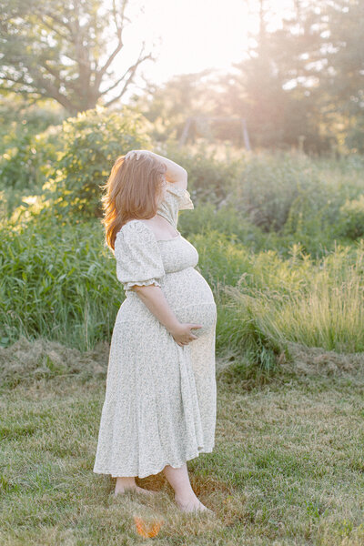 A pregnant woman is standing in a field while holding her hair and looking at the sunset