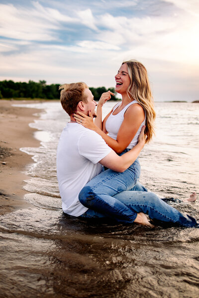 Joyful couple sits straddled on the shores of the beach, their laughter echoing against the sound of the crashing  waves