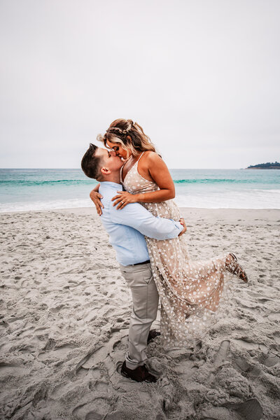 A groom lovingly lifts his bride as they share a passionate kiss on a sandy beach, with calm turquoise ocean waves gently rolling in behind them under an overcast sky.