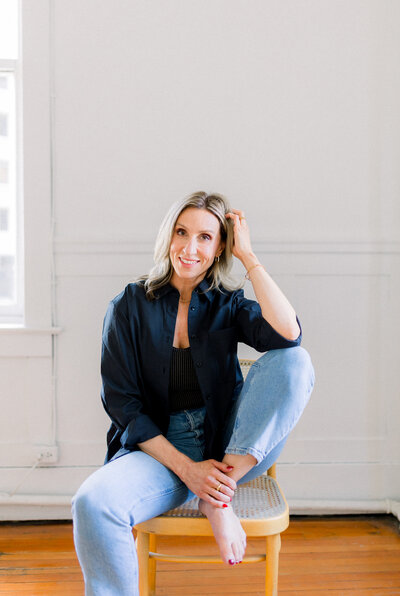 Jennifer Chabot, a Calgary based wedding, brand and lifestyle photographer sitting sitting on a chair with her leg up smiling at camera.