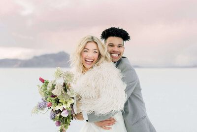 A groom holds his bride around the waist while at the Bonneville Salt Flats at sunset, on their wedding day. 