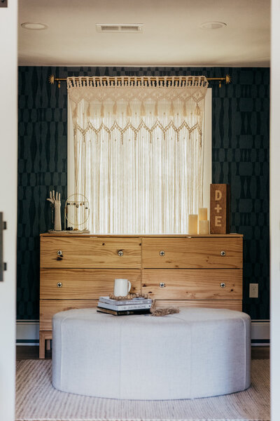 A dresser an ottoman in front of a window with curtains and a tribal pattern wallpaper on the walls representing a colorful interior design by Winsome Home Interiors.