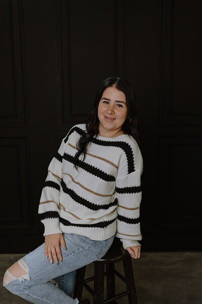 A photo of Britt sitting on a stool while slightly leaning back and smiling at the camera.