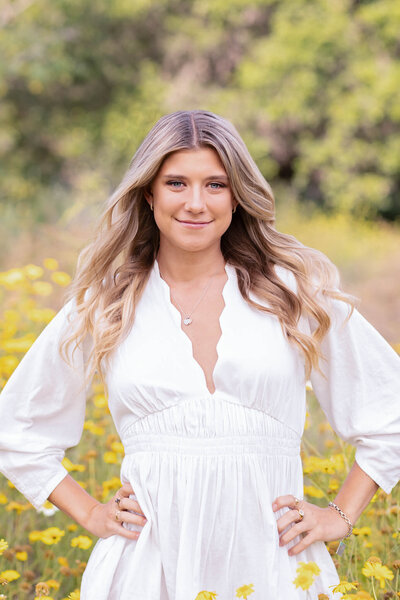 A blonde young woman with long hair wearing a white, long sleeve dress smiling at the camera in a field of yellow flowers. Captured at Northwest Open Space in San Juan Capistrano.