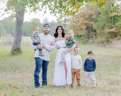 A mom and dad stand in a field holding two toddlers with 2 kids standing with them for a Little Rock family Photographer