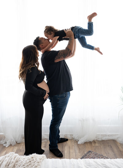engaged couple with dad throwing toddler up in the air infront if a window in a photography studio