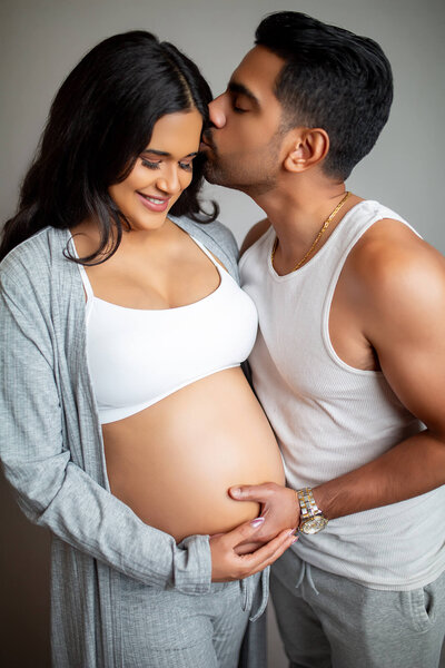 Man kissing wife's head while holding pregnant belly during studio maternity session in Tampa, FL