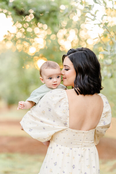 A mother stands in a orchard dressed in a pale yellow dress holding her baby dressed in a pale green jumper as she gently sniffs his head photographed by Bay Area Family Photographer, Light Livin Photography.