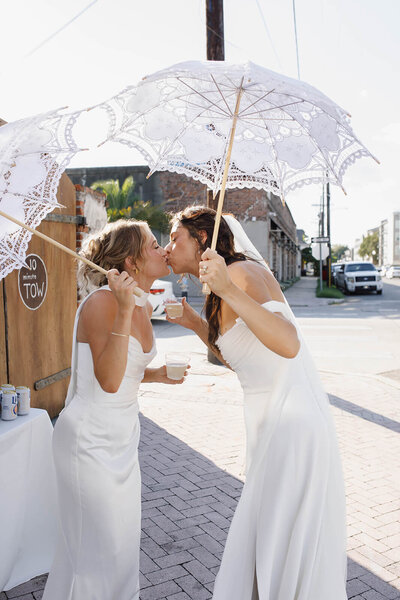 A photo of two brides kissing as they celebrate their marriage holding umbrellas in white dresses