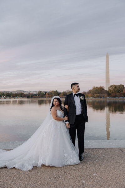 Couple in wedding attire stands in front of the water and Washington Monument on their wedding day.