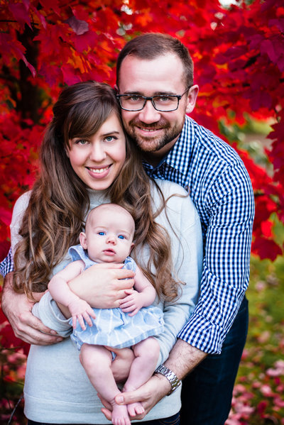 Family in blue hold newborn in front of red fall foliage