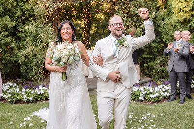 Joyful bride and groom walk up the aisle, celebrating their garden ceremony surrounded by flowers and twinkling lights.