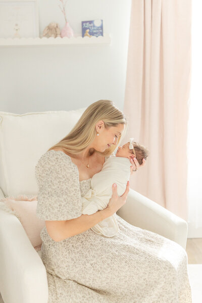 In a cozy home nursery, a mother lovingly gazes at her swaddled newborn as she cradles the baby in her arms. The warm, soft lighting creates a serene atmosphere, highlighting the tender moment between mother and child