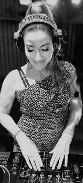 Female DJ wearing headphones and a one-shoulder patterned dress smiling while working the decks at an event.