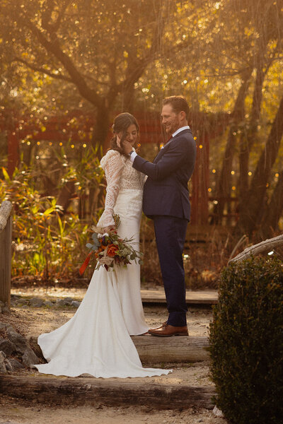 Groom reaching out to his bride who is staring down at her wedding flowers in a garden.