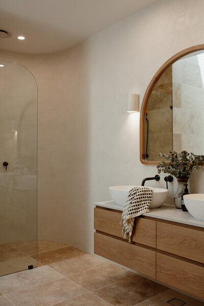 Modern bathroom with a large round mirror, twin vessel sinks, and a wooden vanity next to a glass shower door, showcasing bespoke interior design in Newcastle NSW.