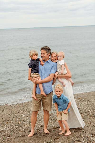 Candid family of four standing by the water during a beach mini session at Sherwood Island in Westport, CT — dad looking at son while the other smiles at the camera