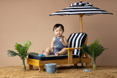 Smiling baby in a blue romper sitting on a miniature lounge chair under a black and white striped umbrella, playing with sand toys on a beach-themed set by a Dallas milestone photographer.