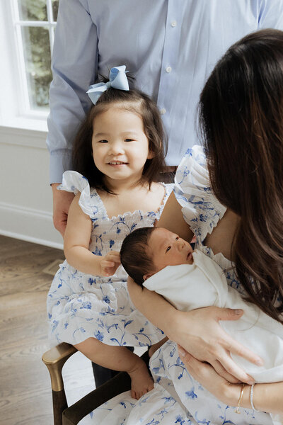 A small child smiling as they sit next to their parent holding a newborn 