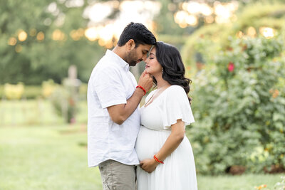 An expecting mother dressed in a white dress, is standing holding her beautiful bump as her husband, who is standing directly in front of her, gently caresses her face, photographed by Tambella Photography