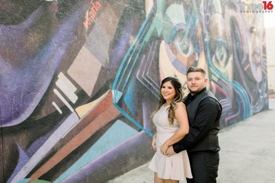 Soon to be Groom holds his fiance from behind as they pose for engagement photos in front of a muraled wall in the Santa Ana Historic District