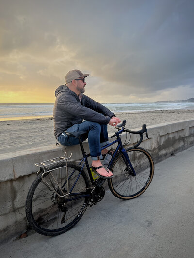 Brian, of Scenic Vows, sits next to his bike  while watching the sunset over the Pacific.