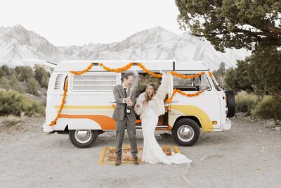 A bride and groom laugh while popping champagne in front of their vintage VW kombi camper while celebrating their wedding in Mammoth Lakes