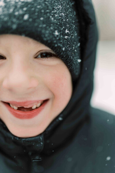 Boy wearing black coat and beanie covered with snowflakes smiles into camera with missing front teeth by Portland Family Photographer Emilie Phillipson Photography. 
